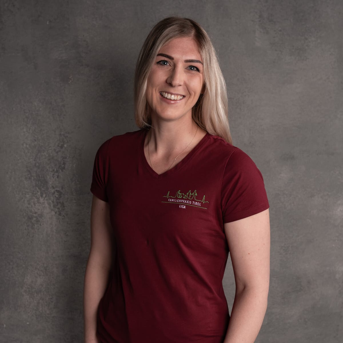 Lisa, infermiera diplomata (DGKP) presso Familienpraxis Tirol, in polo bordeaux con il logo dello studio, sorridente su sfondo grigio.