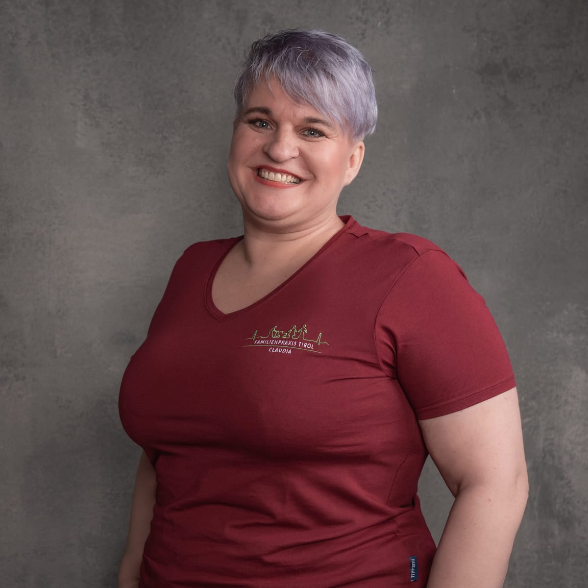 Claudia, assistente infermieristica presso Familienpraxis Tirol, in polo bordeaux con il logo dello studio, sorridente su sfondo grigio.