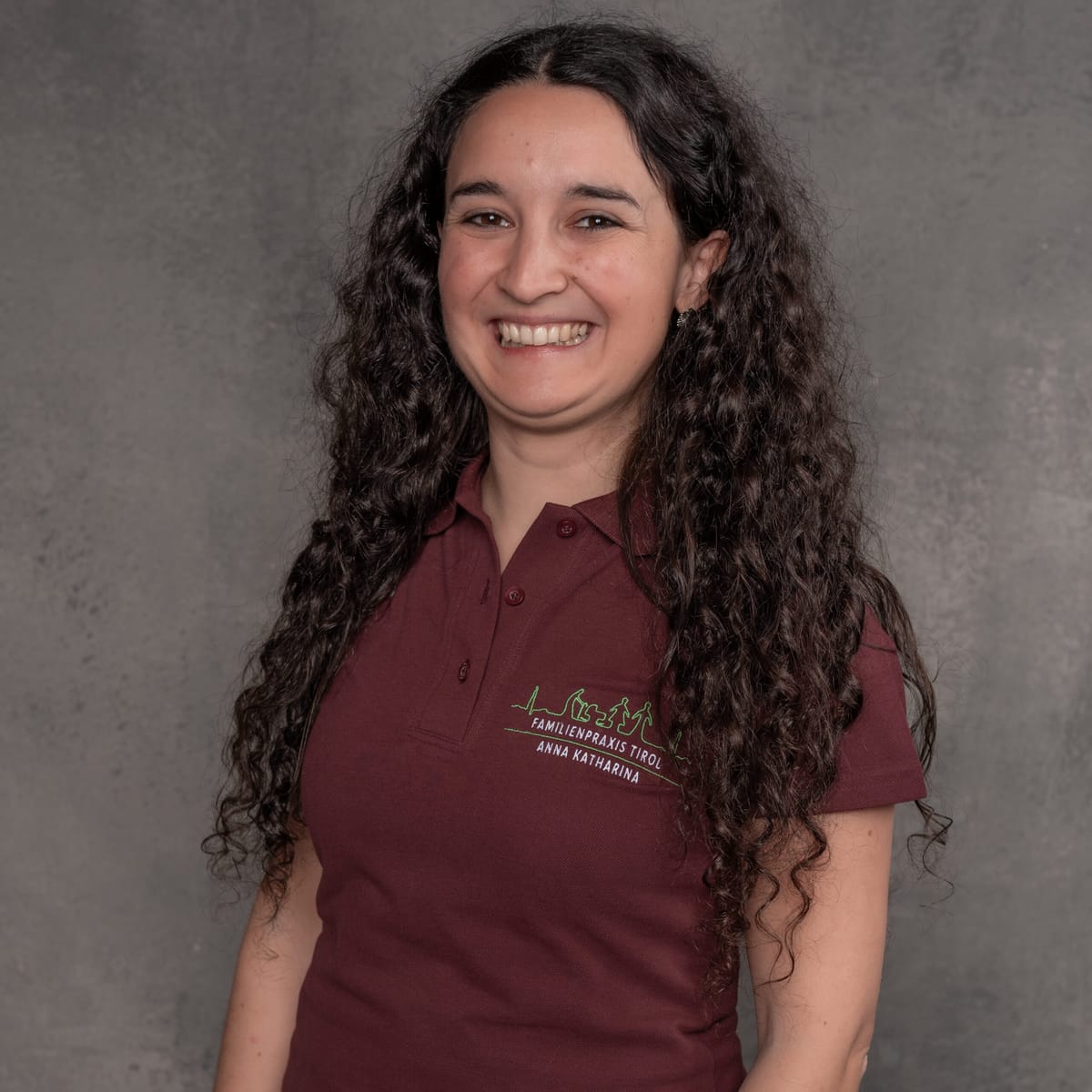 Anna Katharina, responsabile dello studio presso Familienpraxis Tirol, in polo bordeaux con il logo dello studio, sorridente su sfondo grigio.
