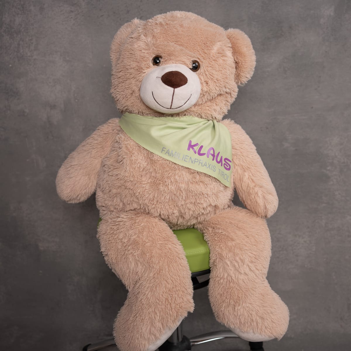Klaus, la mascotte di Familienpraxis Tirol: un grande orsacchiotto beige con foulard e logo dello studio, seduto su una sedia da ufficio verde su sfondo grigio.