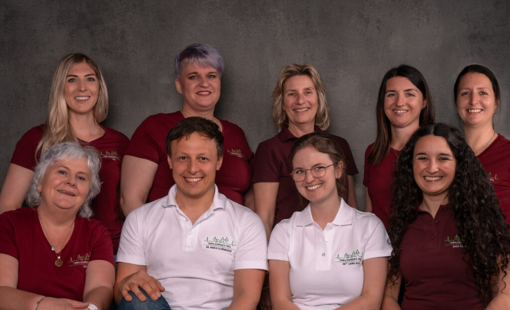 Team der Familienpraxis Tirol, Ihrem Hausarzt in Wörgl, vor grauem Hintergrund, sitzend und stehend gruppiert. In der vorderen Reihe Dr. Marco Schönberger und Dr.in Laura Riepl in weißen Poloshirts, umgeben von den medizinischen und administrativen Mitarbeiterinnen der Praxis in bordeauxfarbenen Shirts.