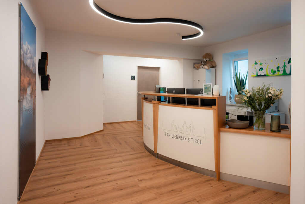 „Empfangsbereich der Familienpraxis Tirol mit modernem Holzdesign, geschwungener Deckenbeleuchtung und freundlich gestalteter Anmeldung mit Praxislogo.“