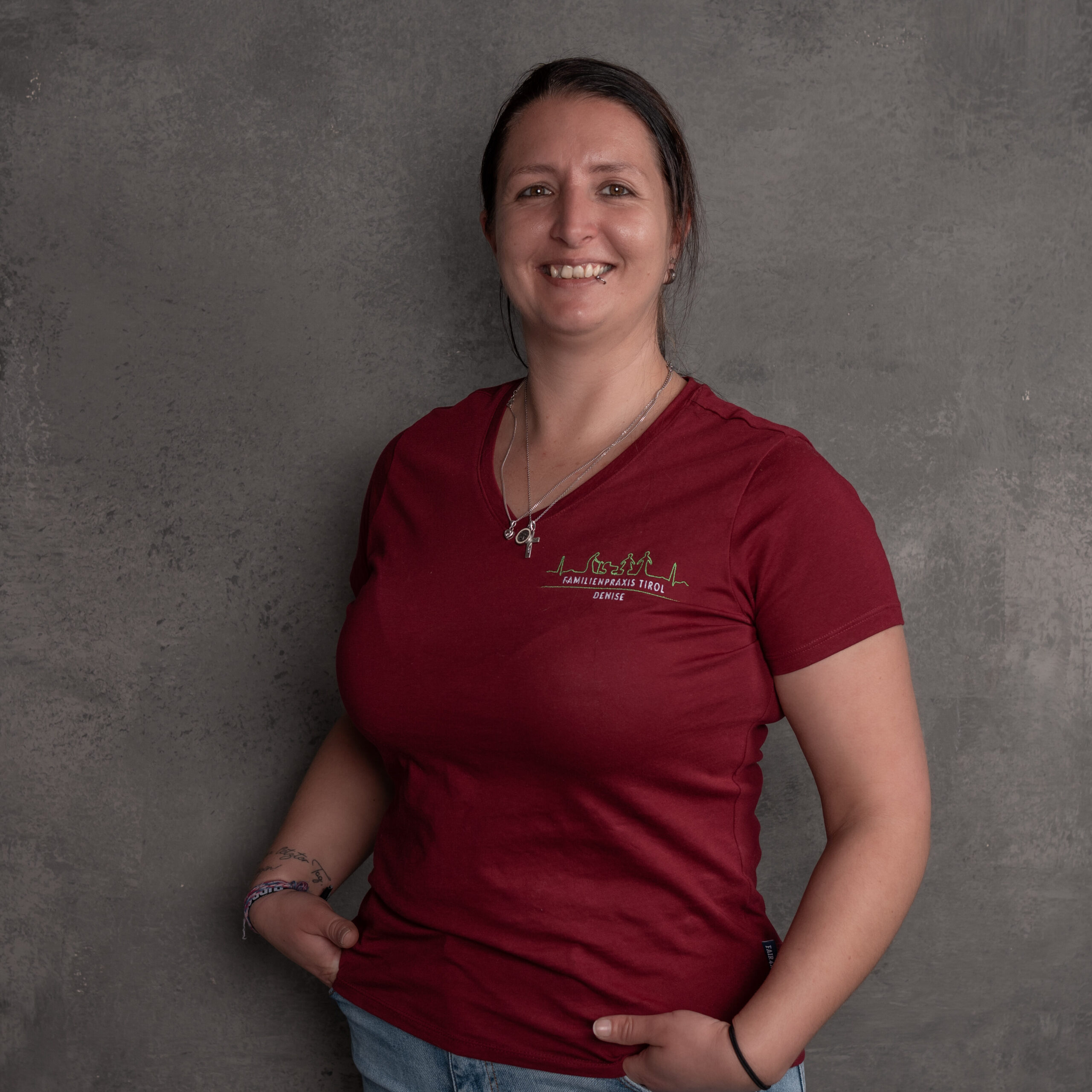 Denise, Reinigungs-, Hygiene und Sterilisationskraft in der Familienpraxis Tirol, im bordeauxfarbenen Poloshirt mit Praxislogo, lächelnd vor grauem Hintergrund.