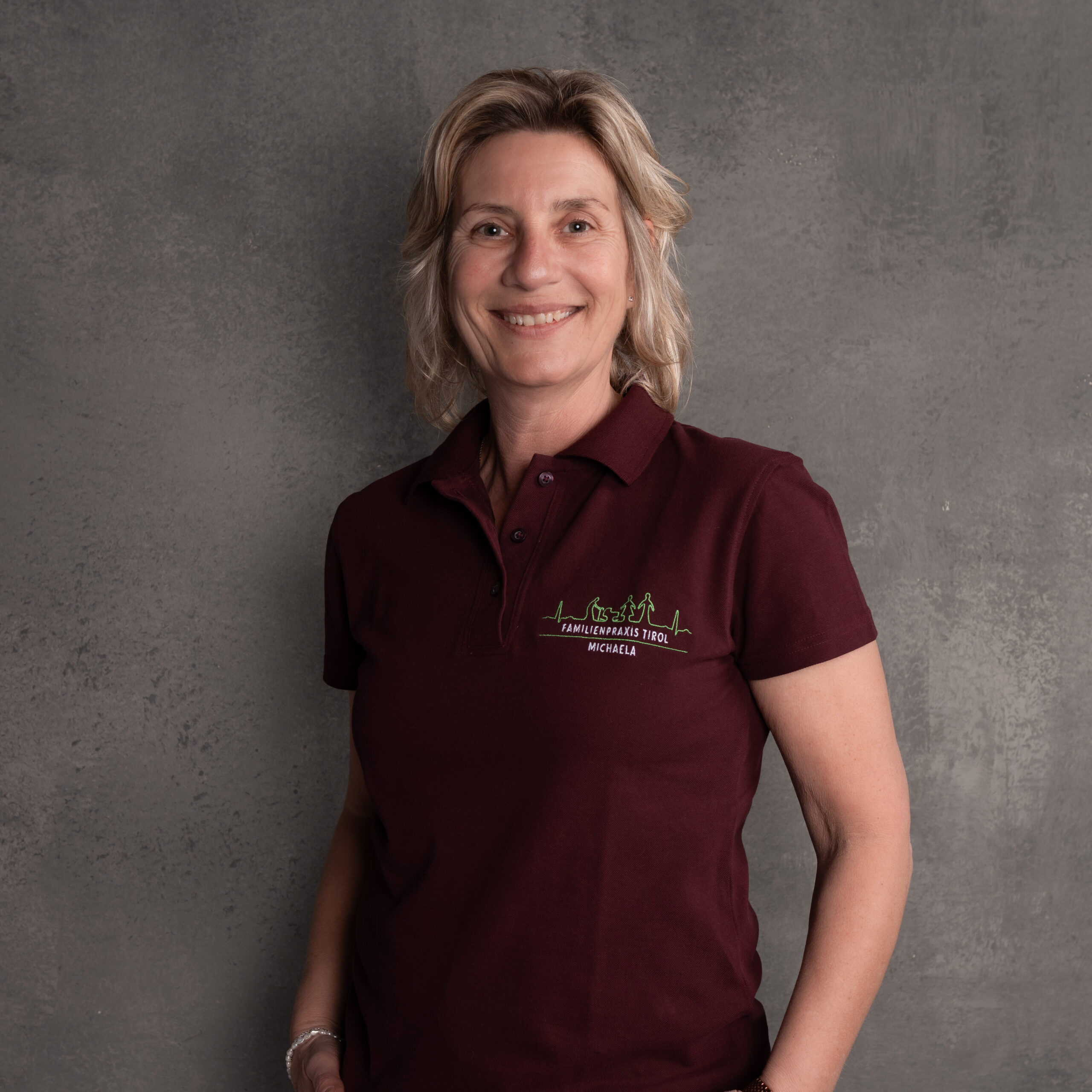 Michaela, Administrativkraft in der Familienpraxis Tirol, im bordeauxfarbenen Poloshirt mit Praxislogo, lächelnd vor grauem Hintergrund.