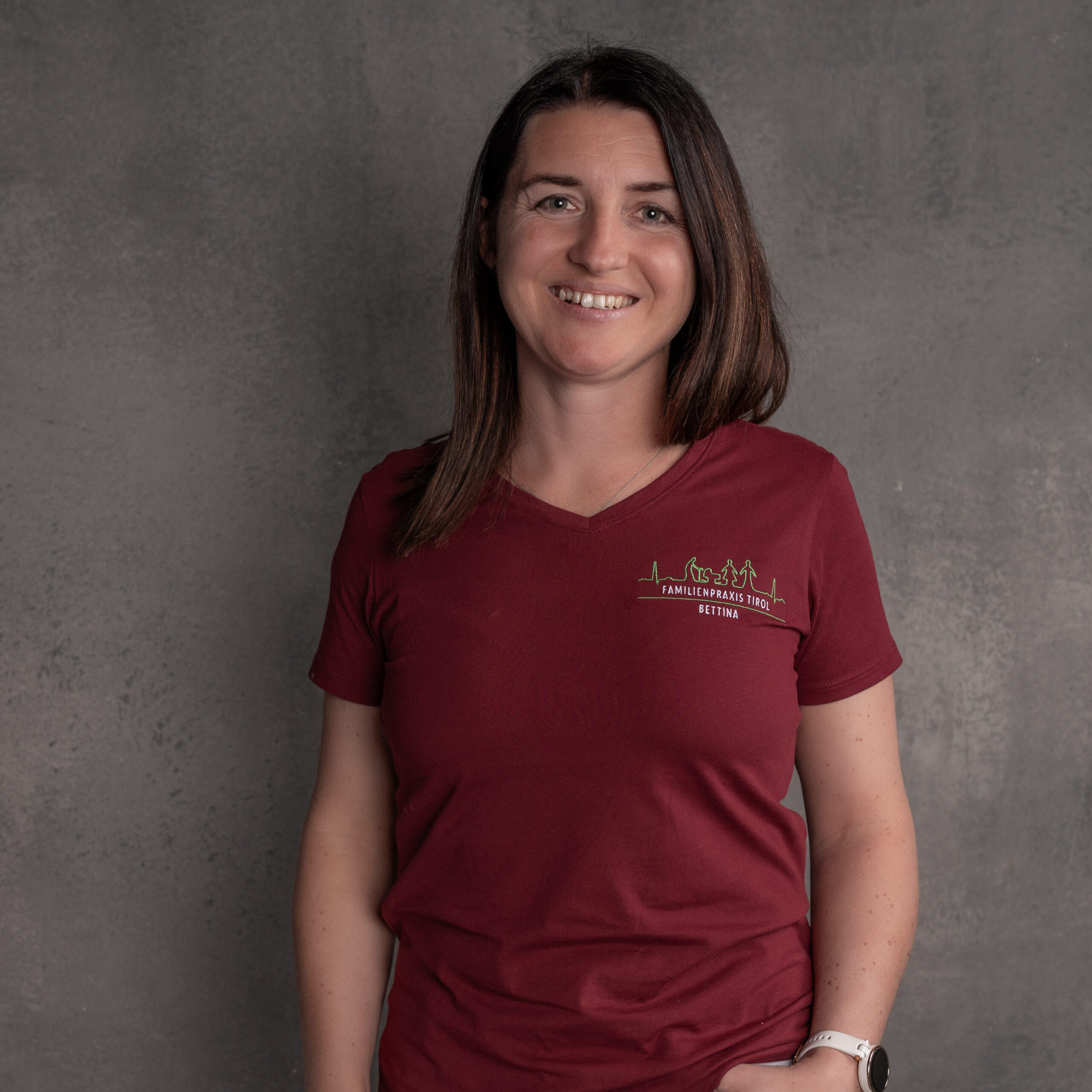 Bettina, Ordinationsassistentin in der Familienpraxis Tirol, im bordeauxfarbenen Poloshirt mit Praxislogo, lächelnd vor grauem Hintergrund.