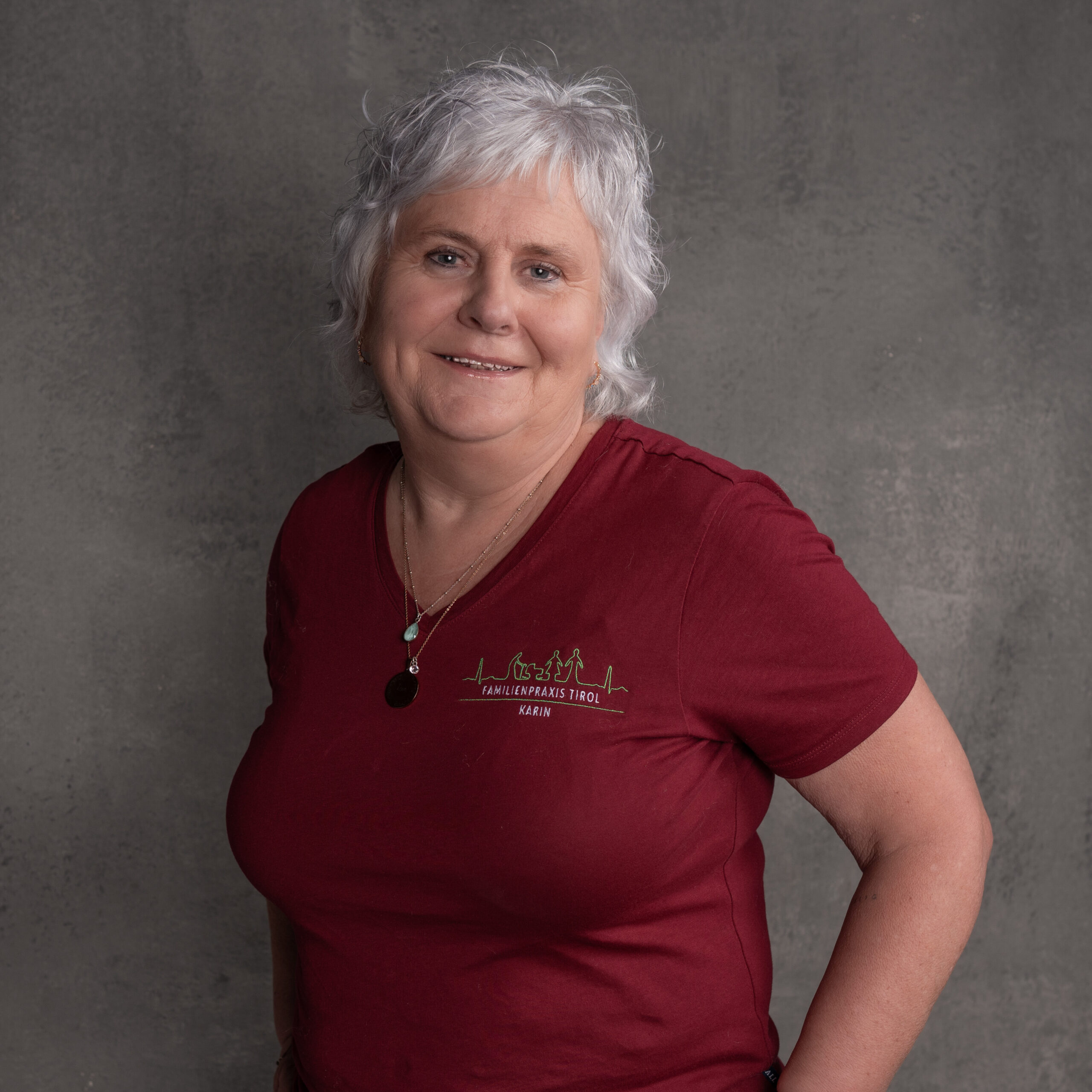 Karin, Ordinationsassistentin in der Familienpraxis Tirol, im bordeauxfarbenen Poloshirt mit Praxislogo, lächelnd vor grauem Hintergrund.