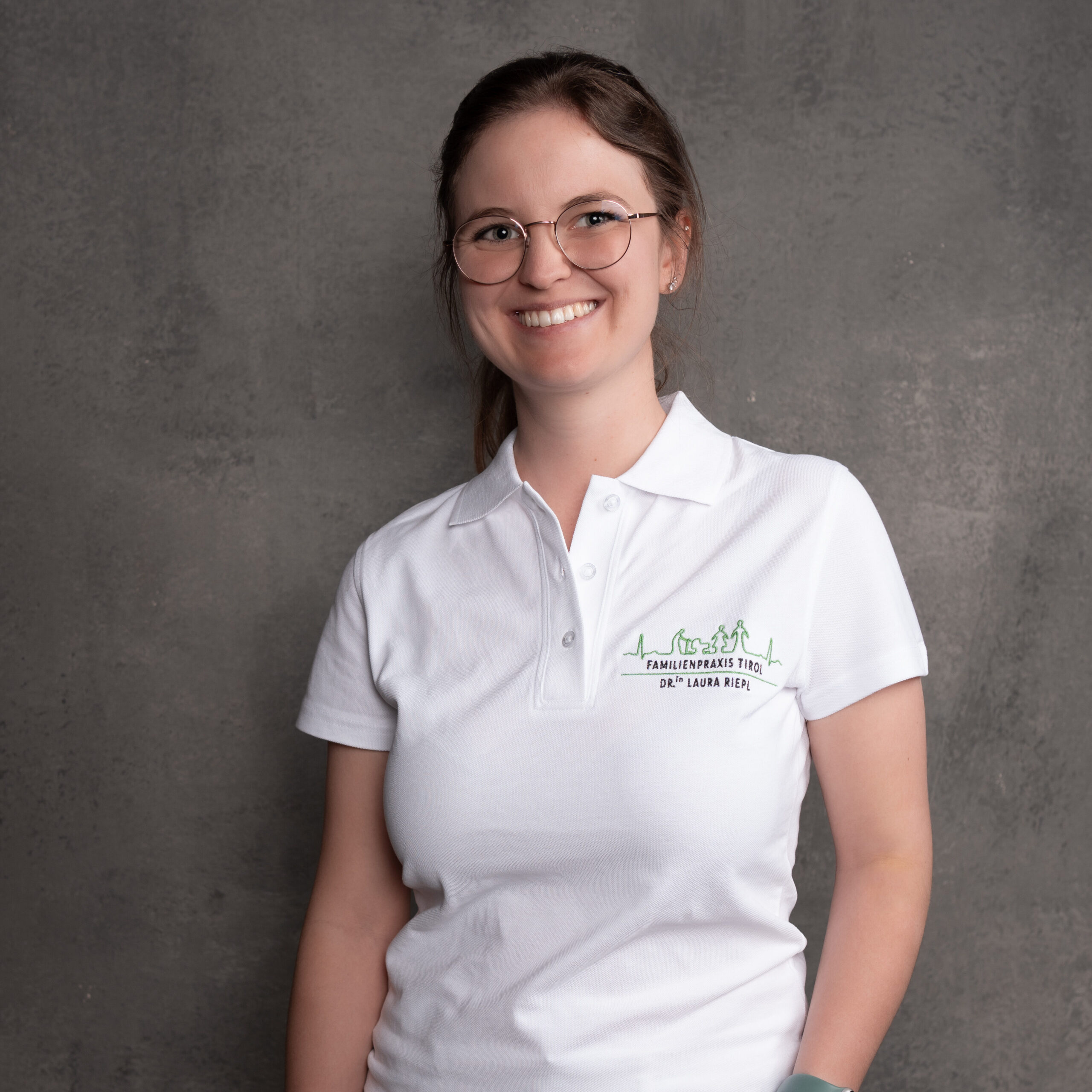 Frau Dr.in Laura Riepl in weißer Praxisbekleidung vor grauem Hintergrund, mit freundlichem Blick in die Kamera.