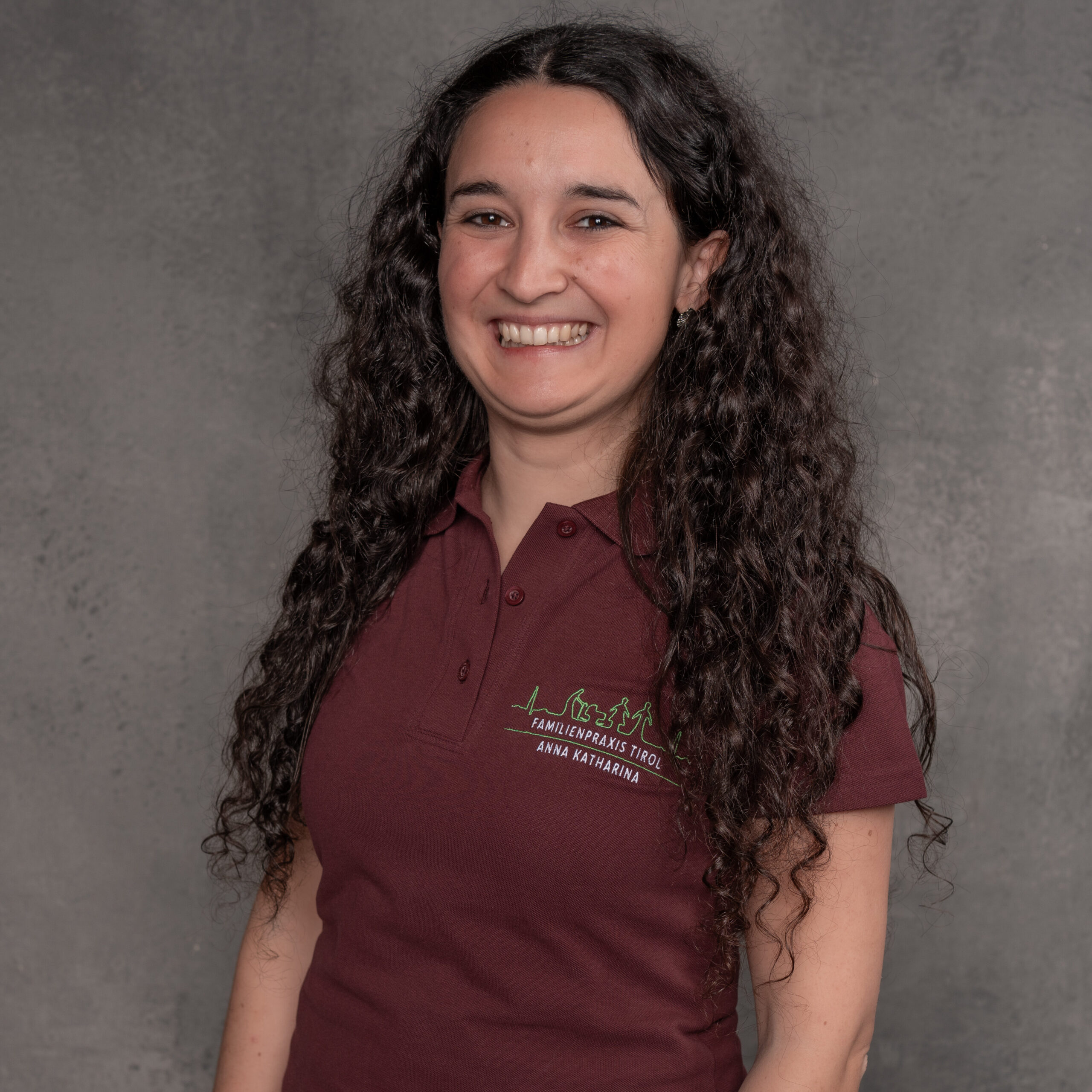 Anna Katharina, Ordinationsmanagerin der Familienpraxis Tirol, im bordeauxfarbenen Poloshirt mit Praxislogo, lächelnd vor grauem Hintergrund.